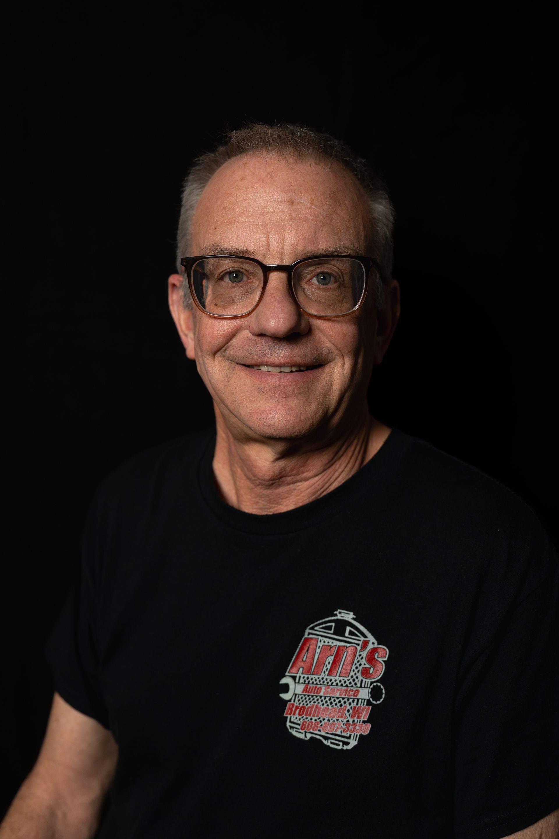 Man with glasses, shaved head, black shirt with logo, smiling against black background.  | Arn's Auto Service