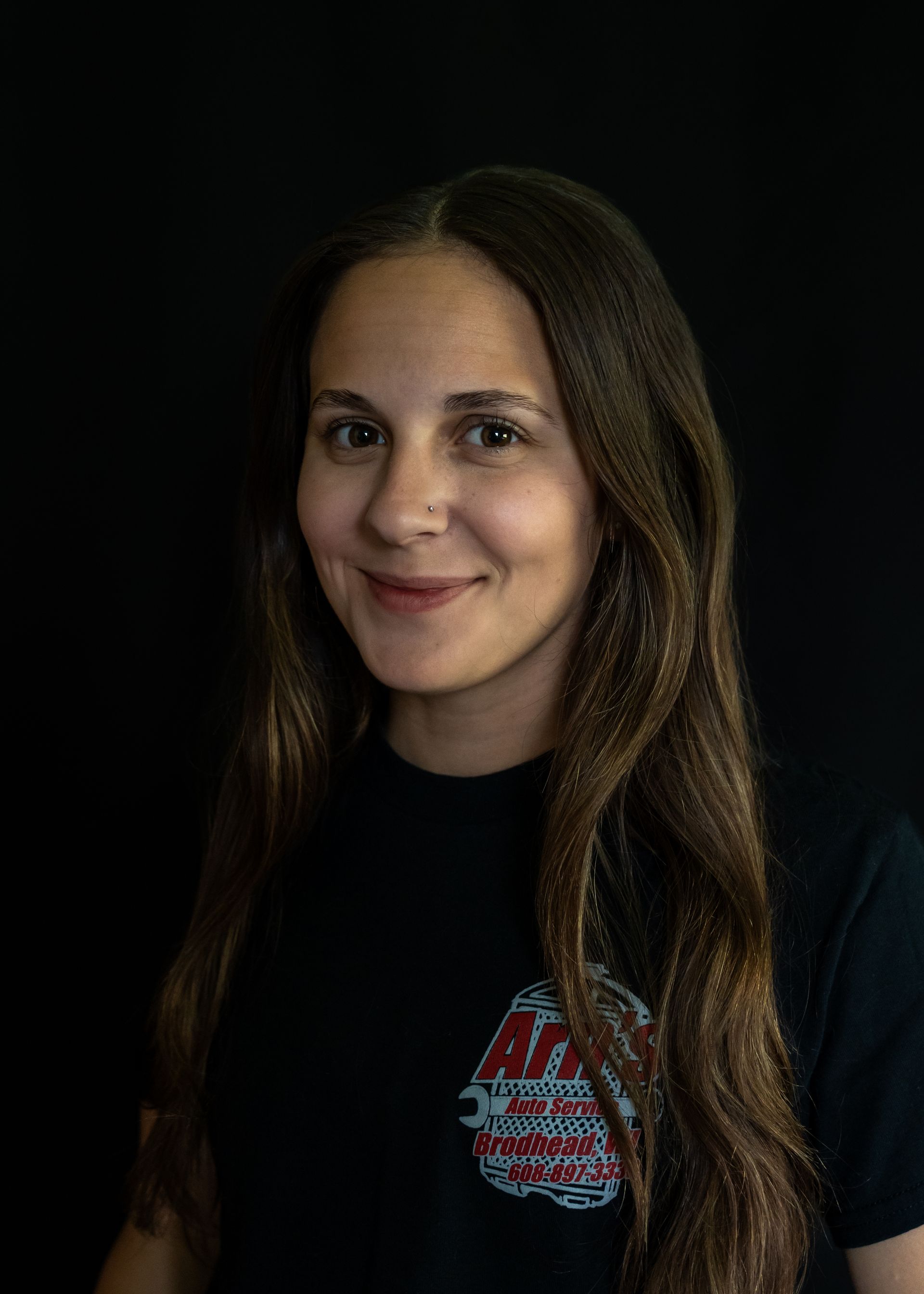 Woman with long brown hair, wearing a black shirt with a logo, smiles at the camera against.  | Arn's Auto Service