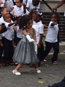 A little girl in a dress and white gloves is dancing in front of a group of children