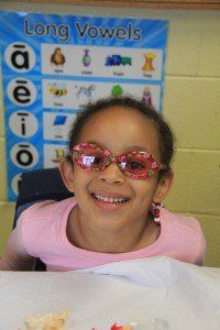 A young girl wearing sunglasses is smiling in front of a long vowels poster