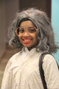 A young girl wearing a gray wig and glasses is smiling.