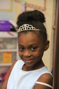 A young girl wearing a tiara and a blue dress is smiling.