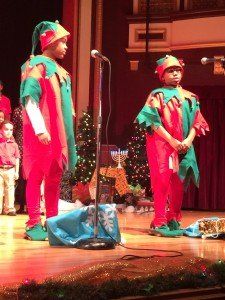 Two children dressed as elves are singing into a microphone