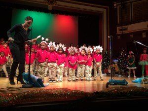 A woman is singing into a microphone in front of a group of children wearing santa hats.