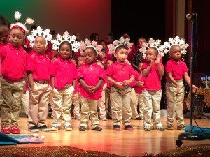 A group of children wearing red shirts and white pants are standing on a stage.