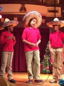 Three children wearing sombrero hats are singing into a microphone on a stage.