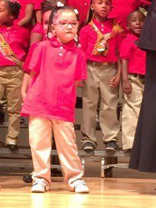 A little girl in a pink shirt is standing in front of a choir.