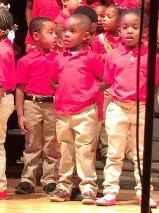 A group of children wearing red shirts and khaki pants are standing on a stage.