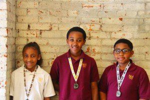 Three children wearing medals are standing in front of a brick wall.