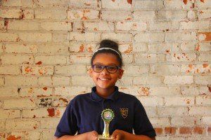 A young girl is holding a trophy in front of a brick wall.