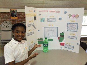 A young girl is holding a bottle of sprite in front of a project board.