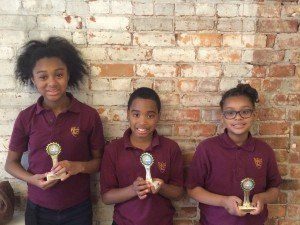 Three children are holding trophies in front of a brick wall.