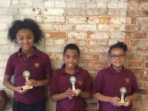 Three young girls are holding trophies in front of a brick wall.