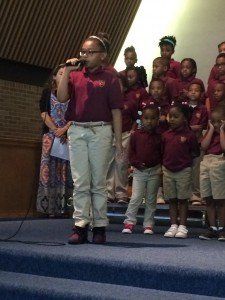 A girl is singing into a microphone in front of a group of children