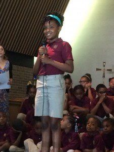 A young girl is speaking into a microphone in front of a group of children