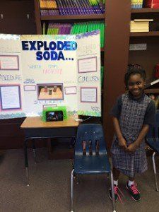 A young girl is standing in front of a poster about exploded soda.