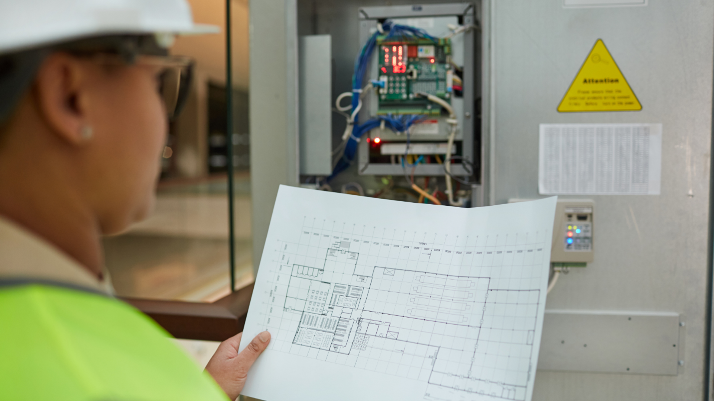 Worker in hard hat and safety vest examining a blueprint in front of an electrical panel.