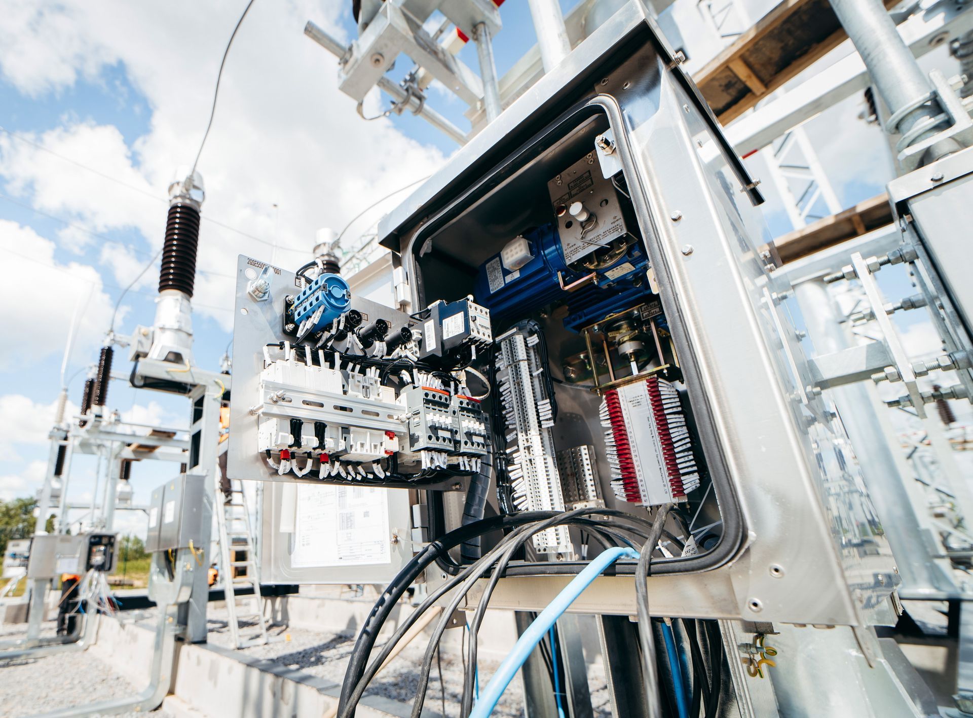 Opened electrical control box at a power substation, blue sky background. Wires and electrical components visible.