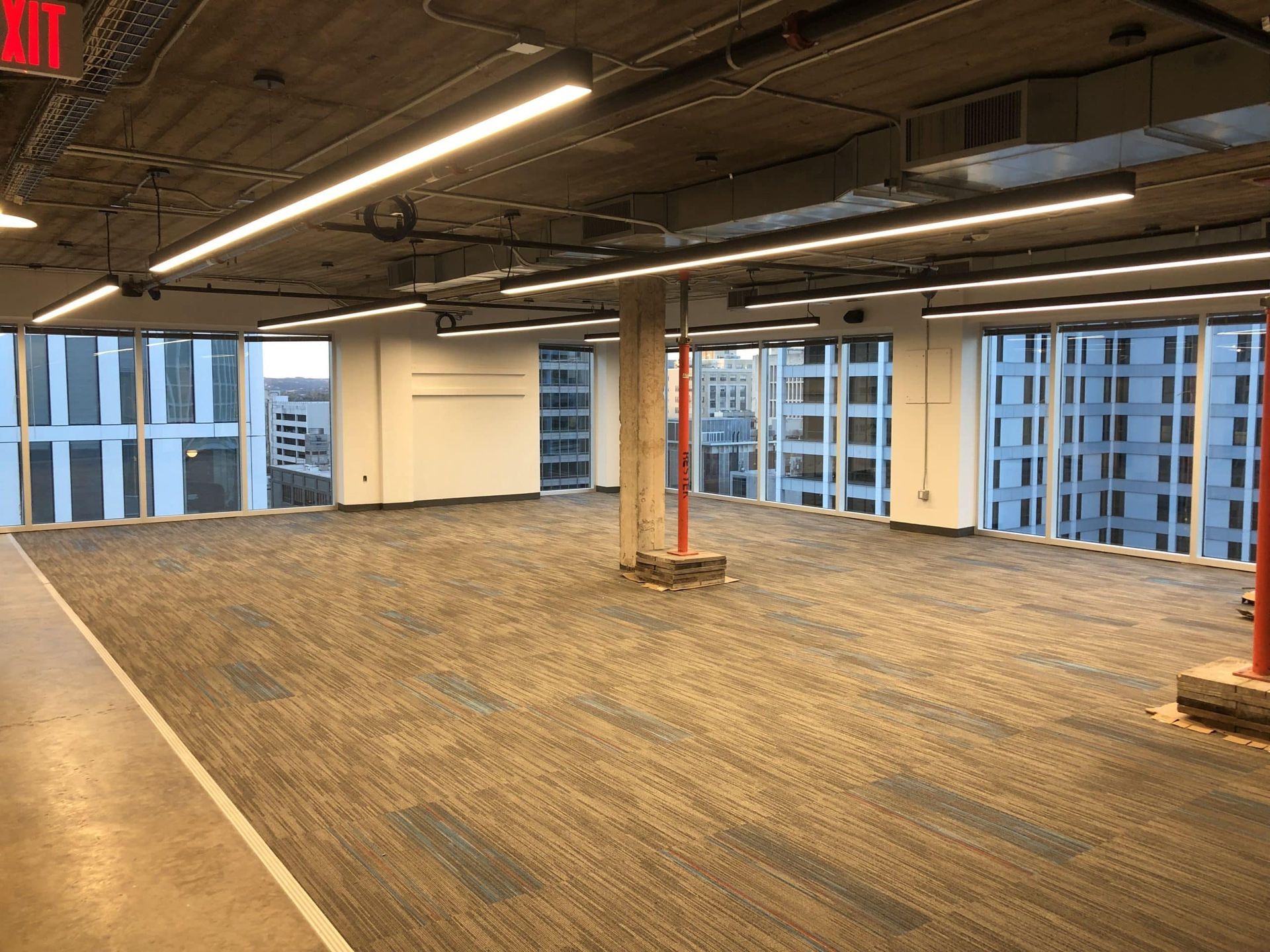 Empty office space with large windows and city views, beige carpet, and exposed ceiling.