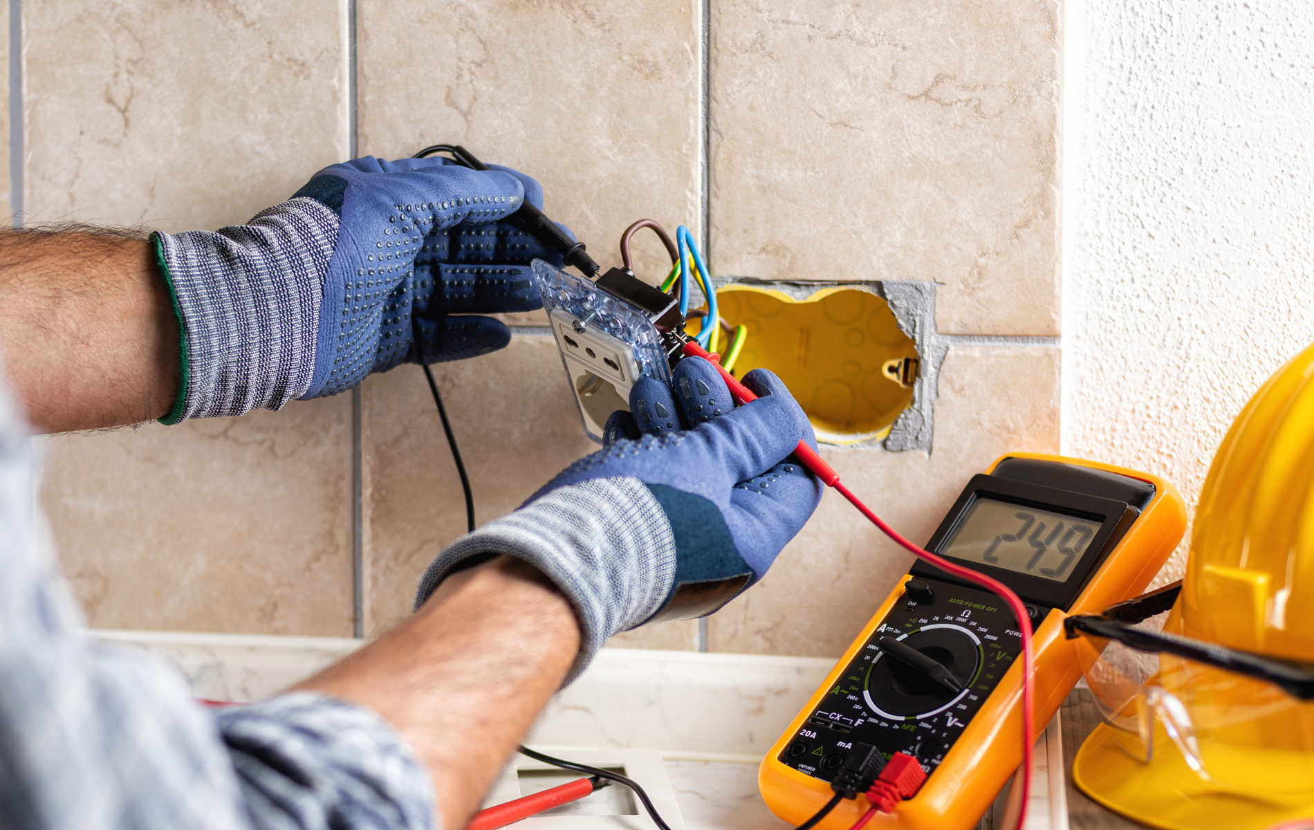 Electrician testing a wall outlet with a multimeter, wearing blue gloves, in a tiled setting.