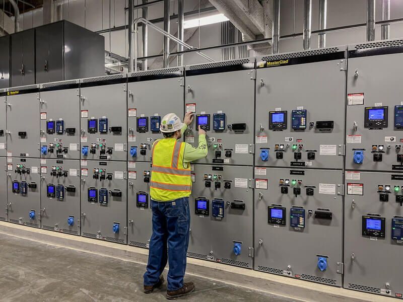 Worker in safety vest at electrical control panel, adjusting equipment. Industrial setting.