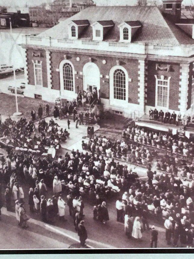 Large crowd gathers outside a red brick building with arched windows and dormers.
