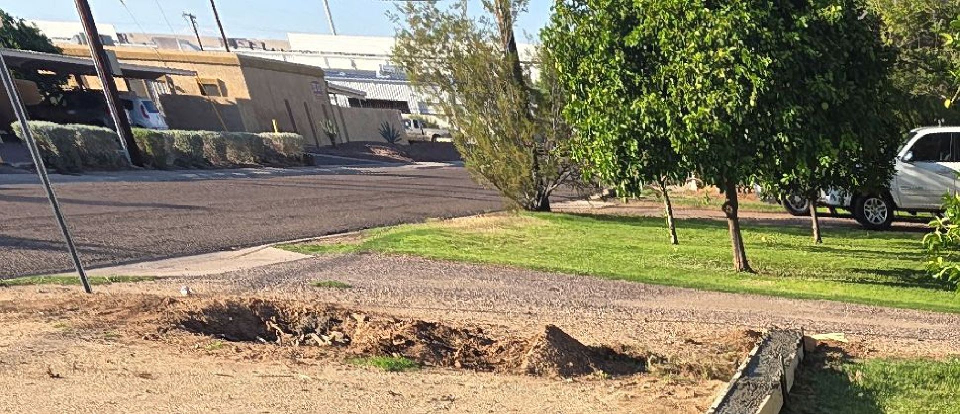A dug-out hole in the dirt, next to a grassy lawn and a paved road with buildings in the background.