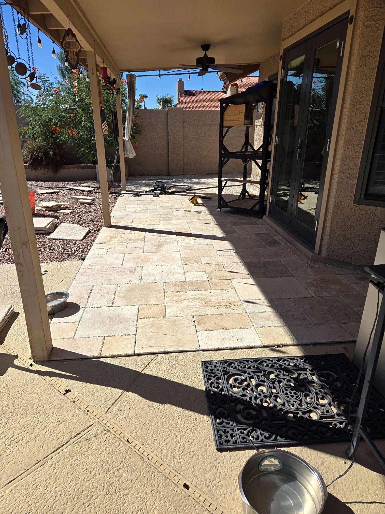 Covered patio with stone tiles. Black door, metal screen, and doormat are visible. Bright sunlight.