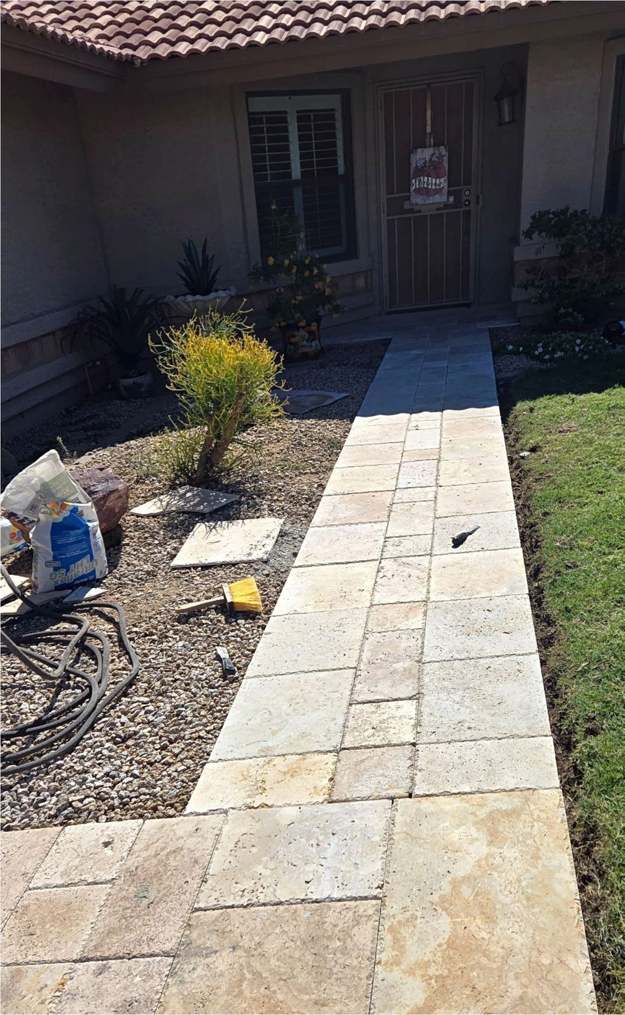 Stone walkway leading to a house entrance. Gravel, plants, and a bag of material on the side.