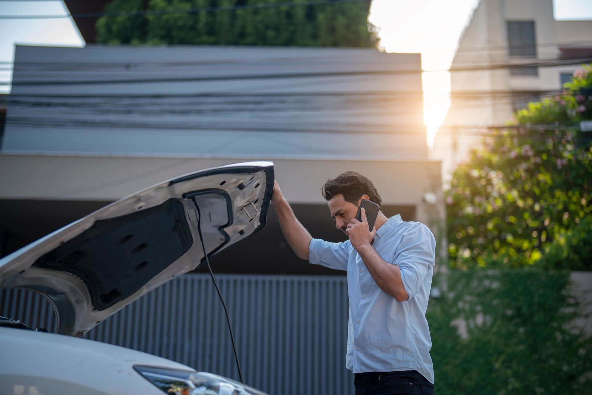 A man is talking on a cell phone while looking under the hood of his broken down car.