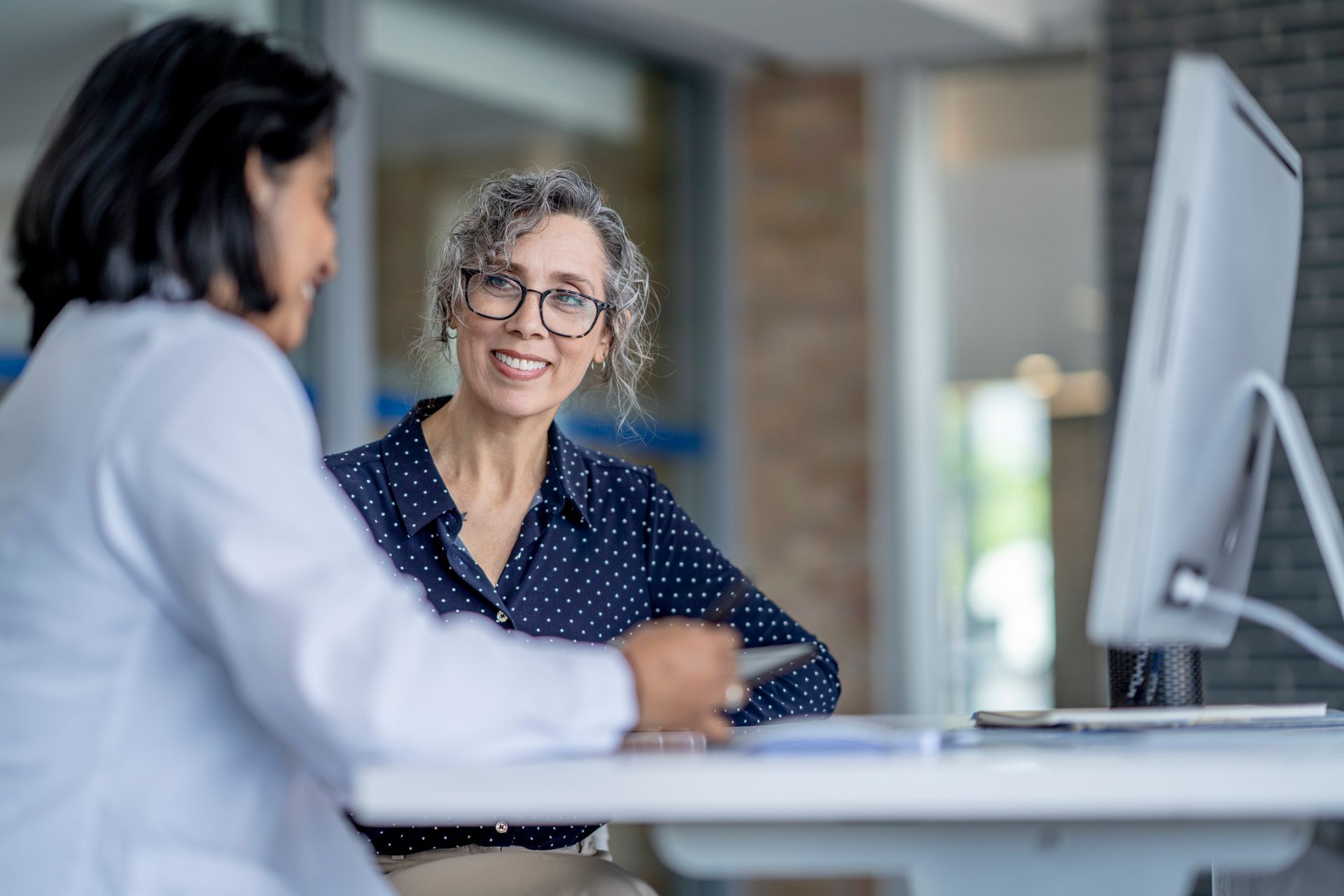 Two women are sitting at a table talking to each other in front of a computer.