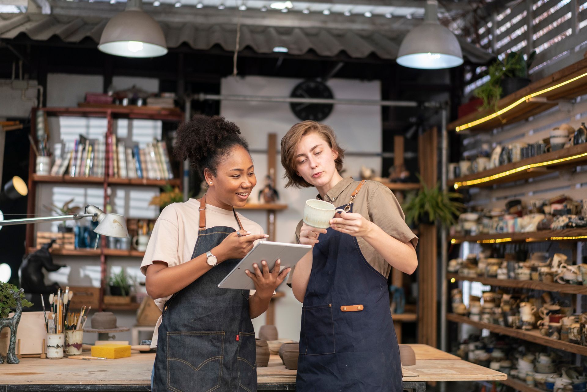 Two women are standing next to each other in a pottery studio looking at a tablet.