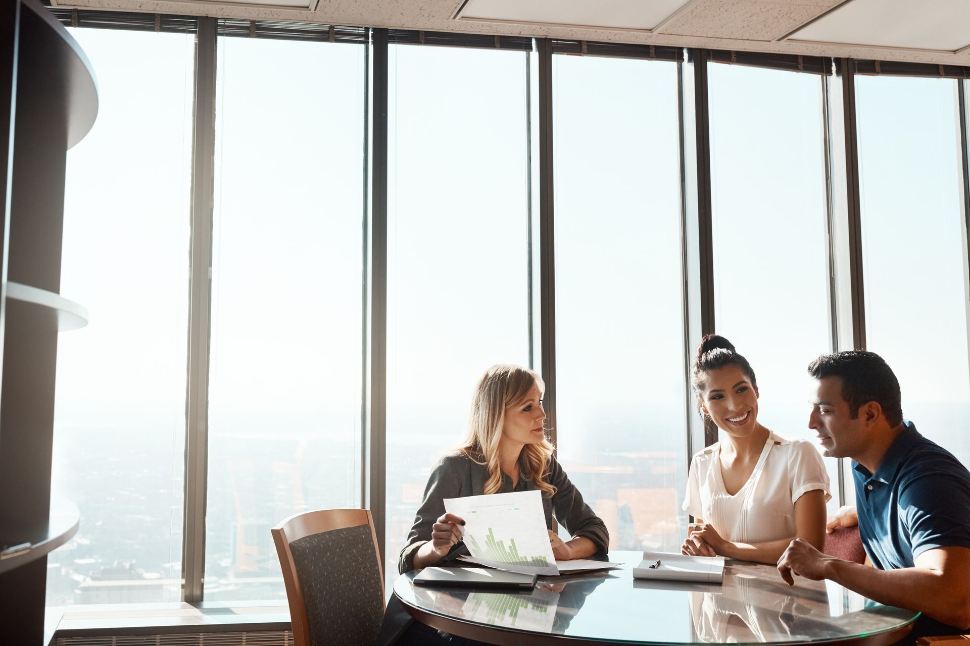 A group of people are sitting at a table in front of a window having a meeting.