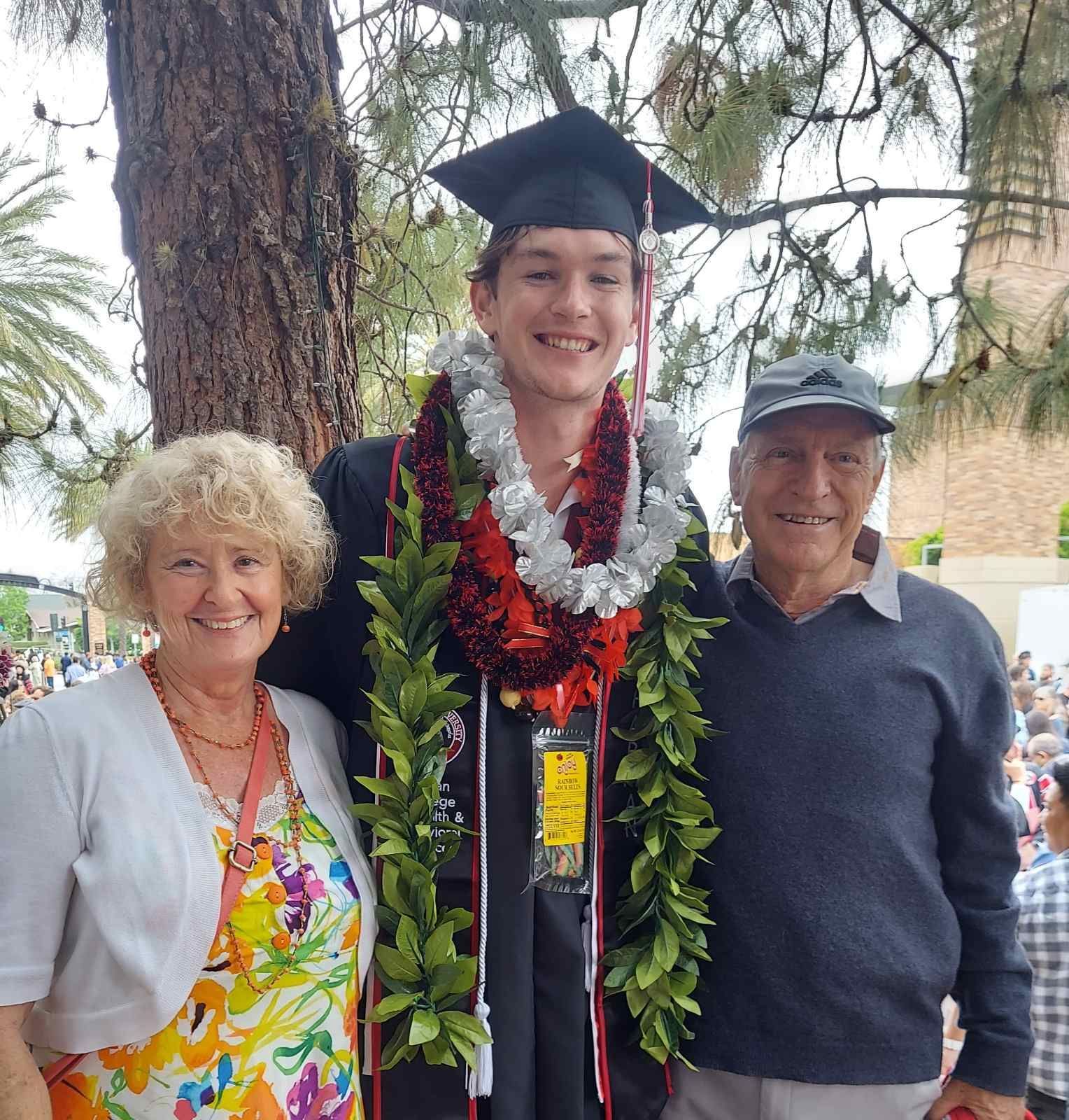 Graduate in cap and gown with leis, smiling between two older adults outdoors
