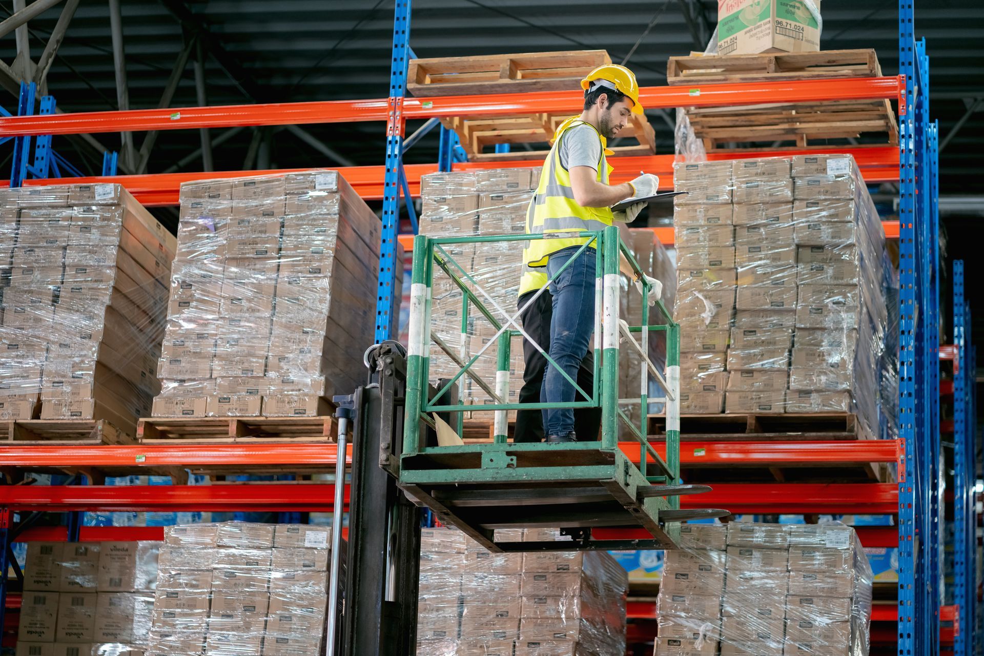 Warehouse worker in lift, inspecting inventory on high shelves; boxes, safety vest, hard hat.
