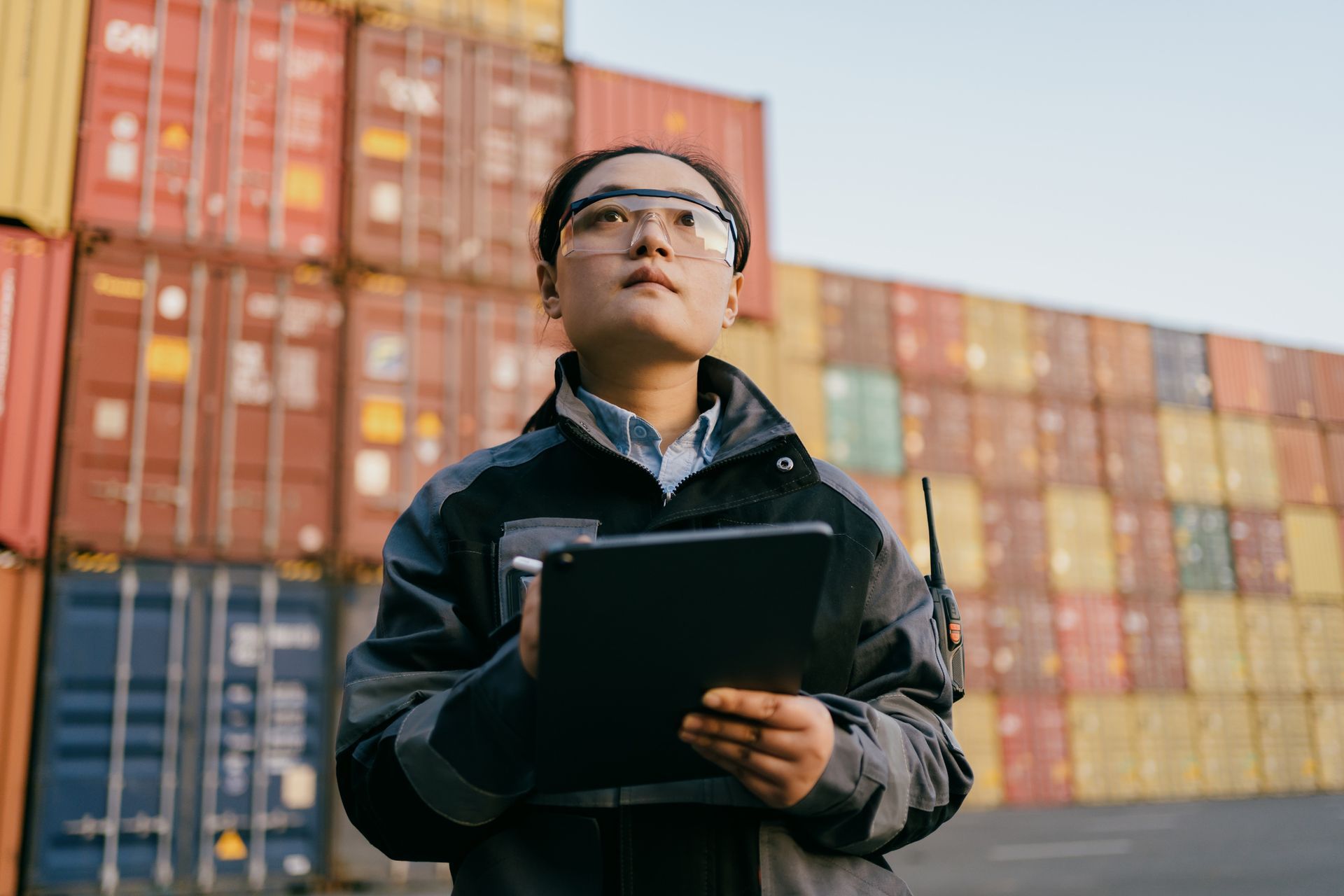 Woman in safety glasses holding a tablet, looking up at shipping containers in a logistics yard.