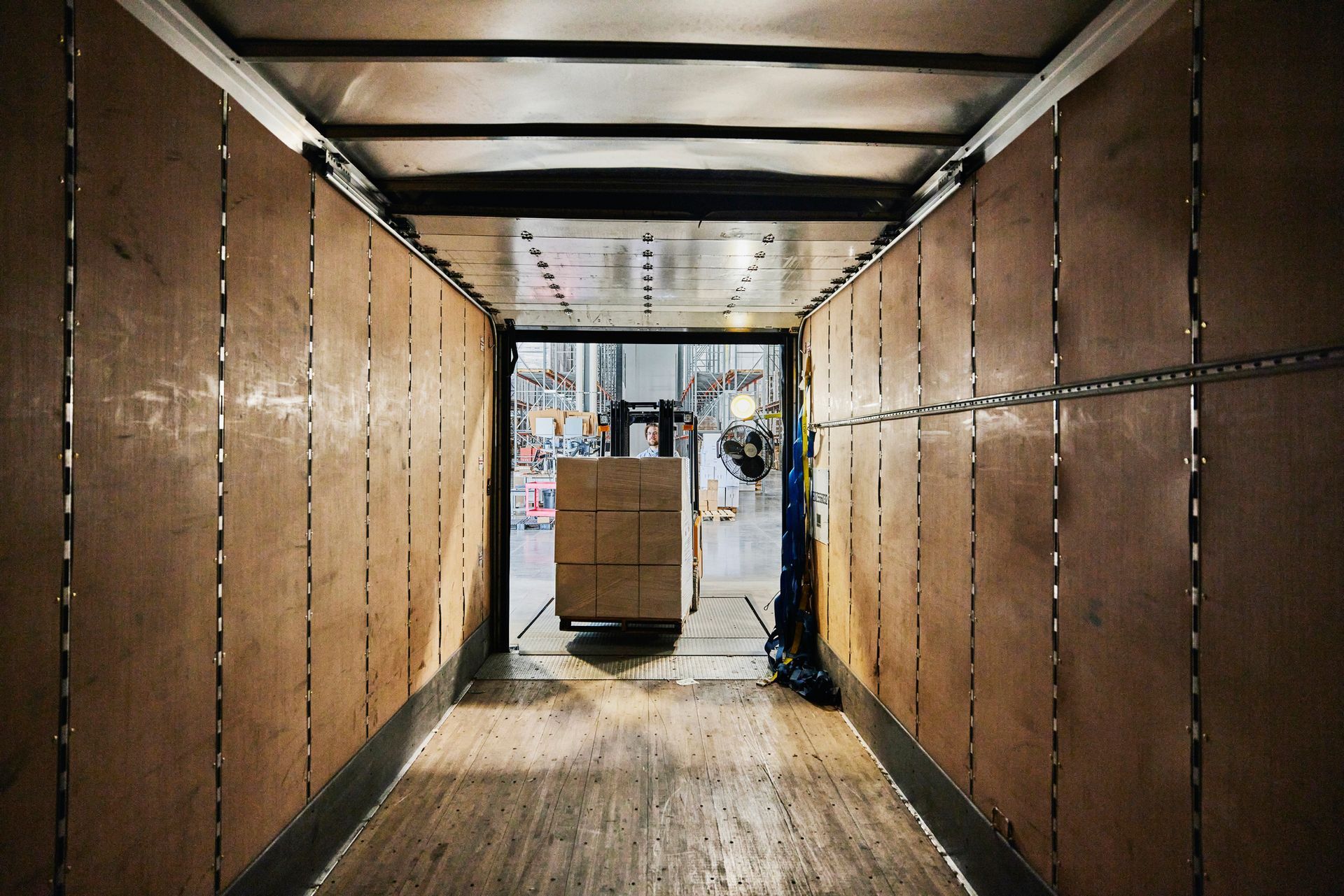Inside a truck, a forklift loads boxes. Brown wood panels line the walls, and the open door reveals a warehouse.