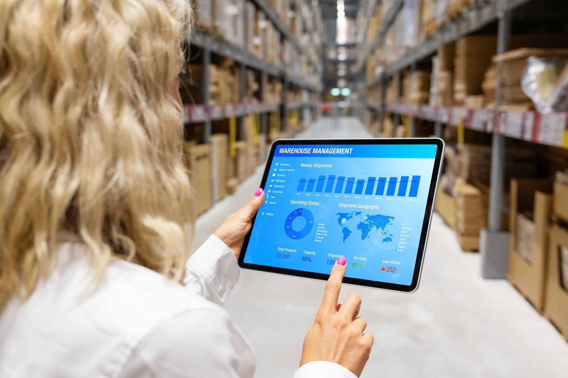 Woman in warehouse holding tablet, reviewing inventory data on a blue screen, pointing at the screen.