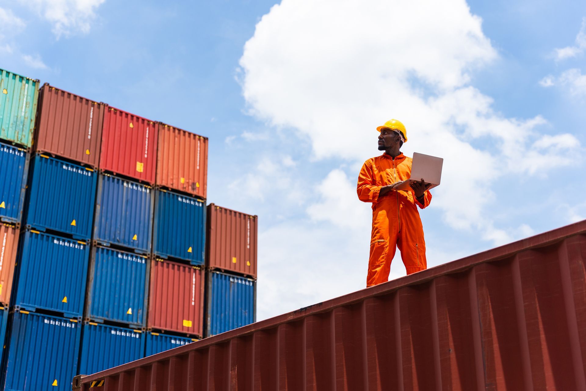 Person in orange jumpsuit and hard hat using laptop on top of shipping containers. Blue sky in background.