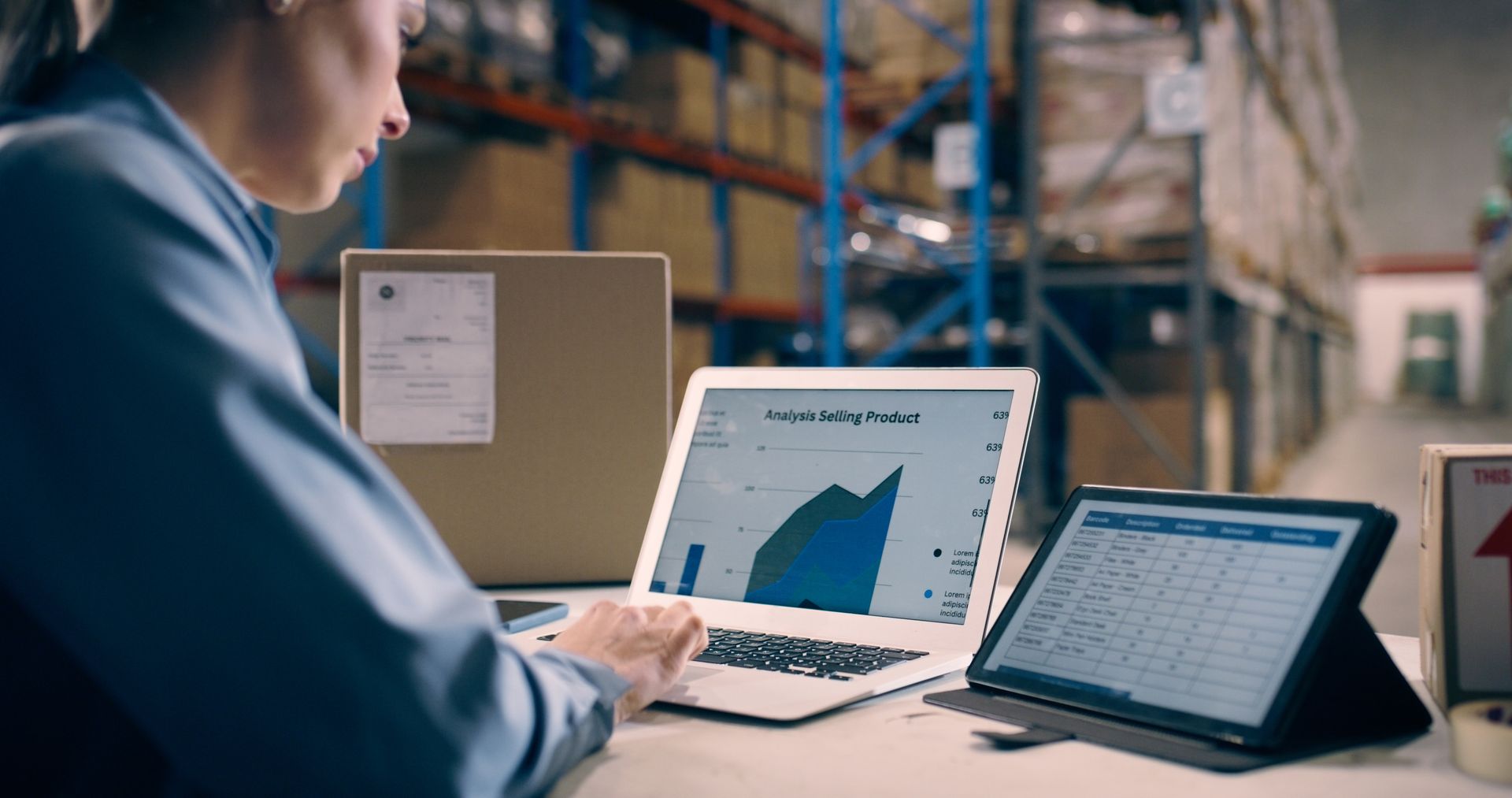 Woman working on a laptop and tablet in a warehouse, analyzing data with boxes in the background.