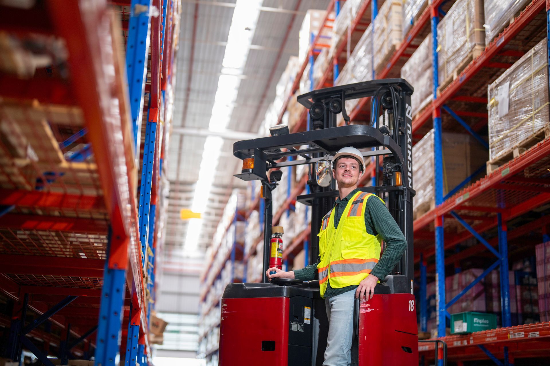 Warehouse worker on a forklift smiling in a storage facility; wearing a hardhat and safety vest.