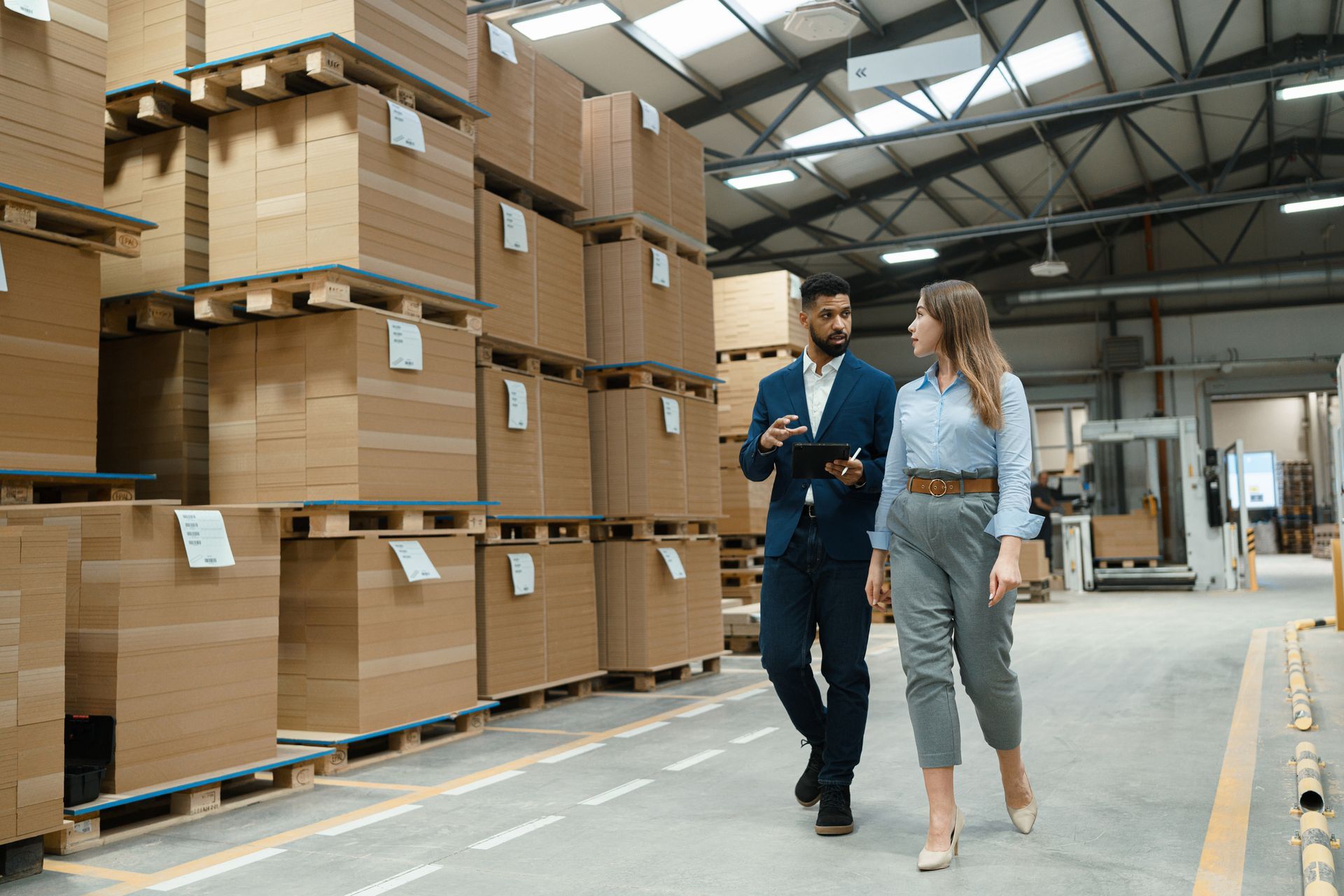 Man and woman walking and talking in a warehouse, surrounded by stacked boxes on pallets.