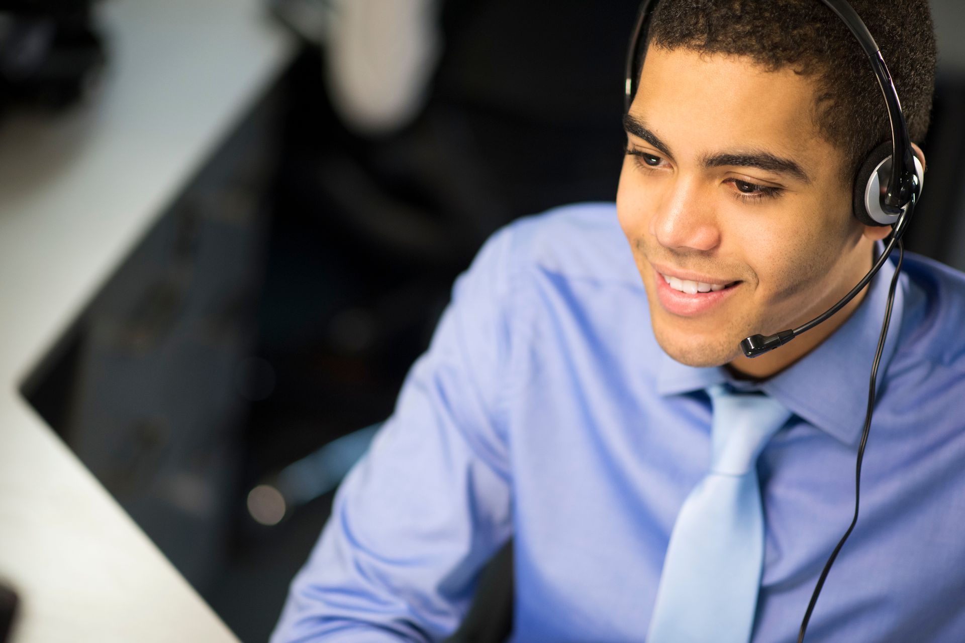 Young man with headset, smiling, wearing a blue shirt and tie in an office setting.