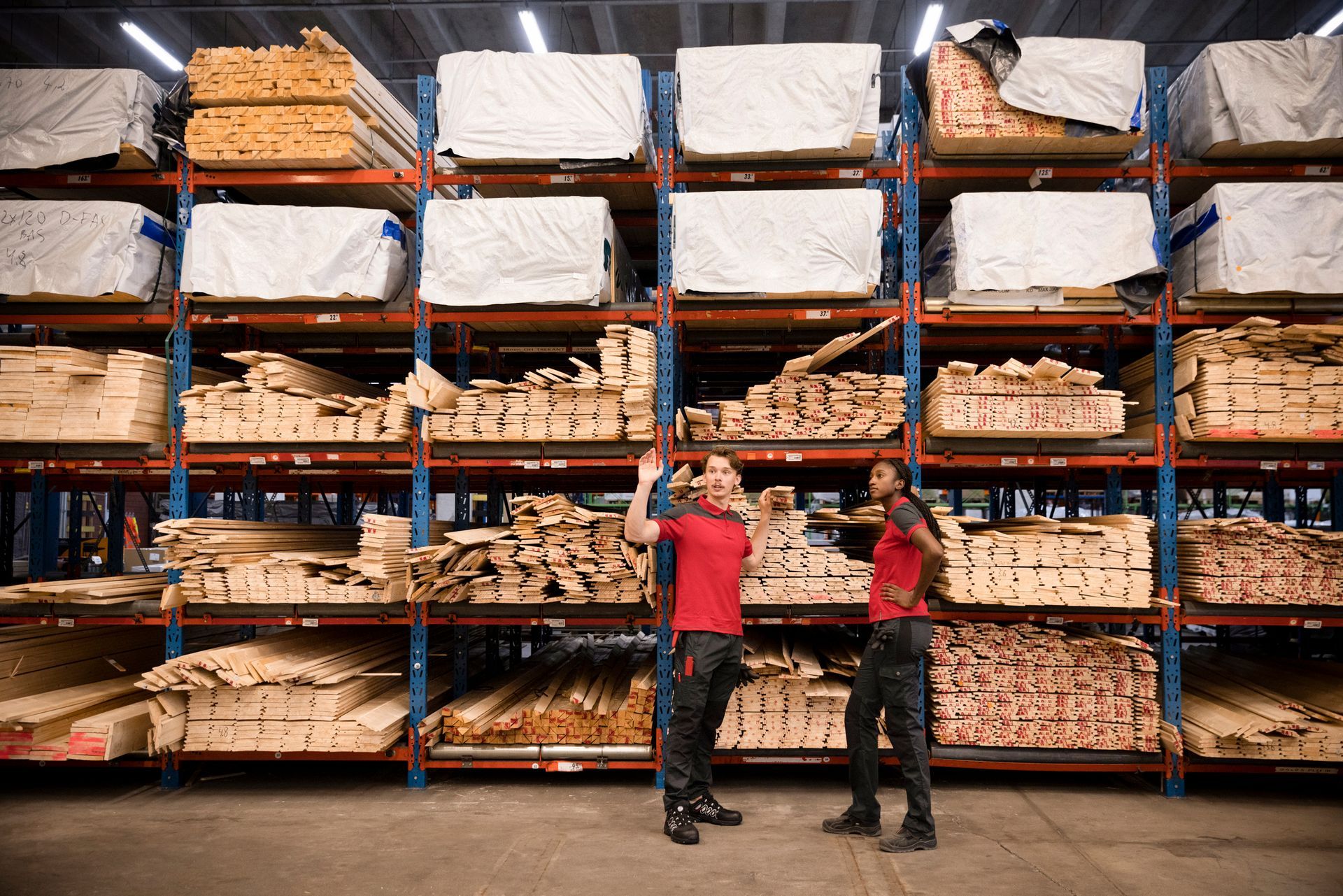 Two workers stand in a lumber warehouse, surrounded by shelves of wood. One gestures, the other looks on.