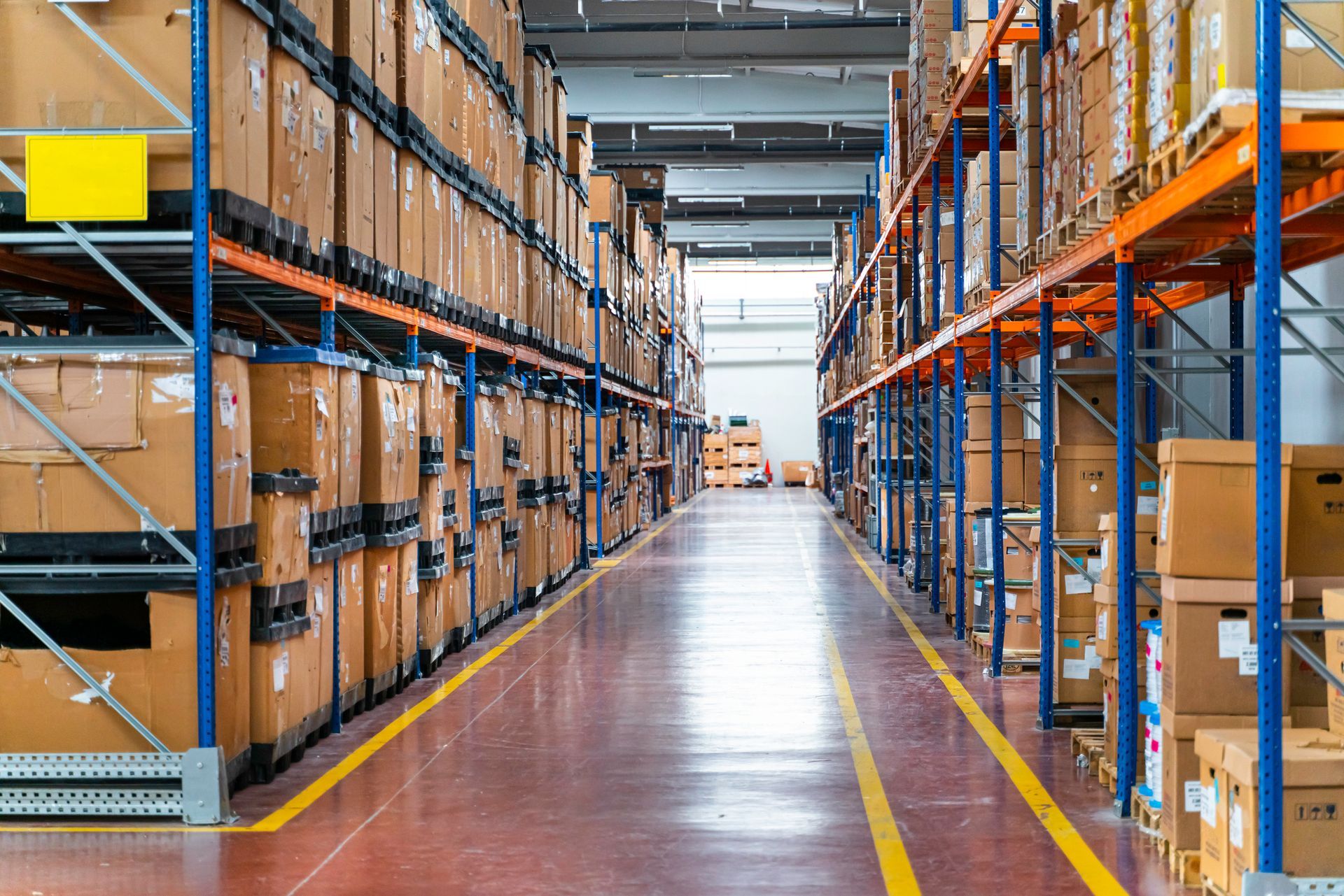 Warehouse aisle with boxes stacked on metal shelving, red floor, yellow lines.