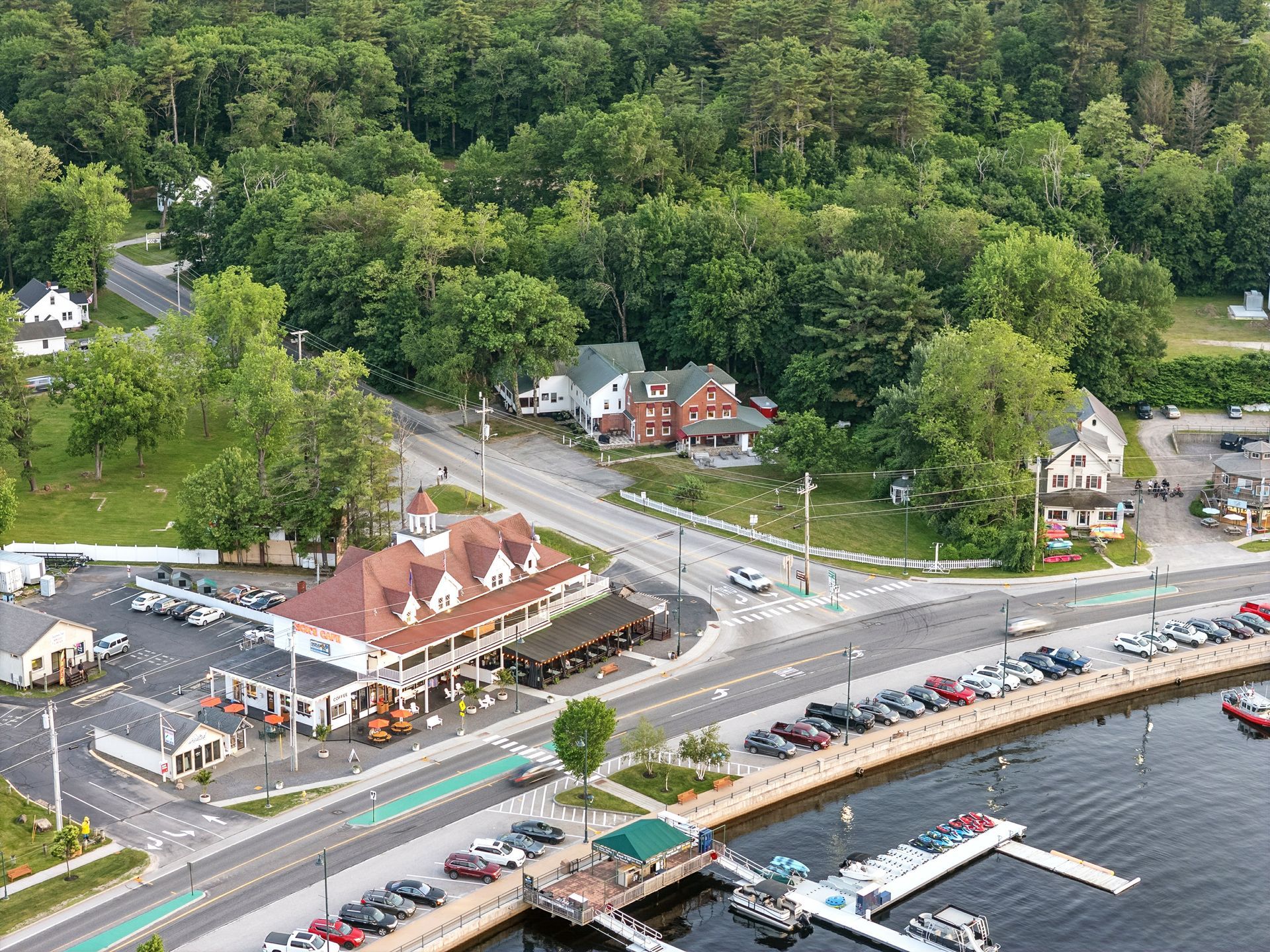 Aerial view of a lakeside town with buildings, roads, parking, boats, and a lush green forest background.