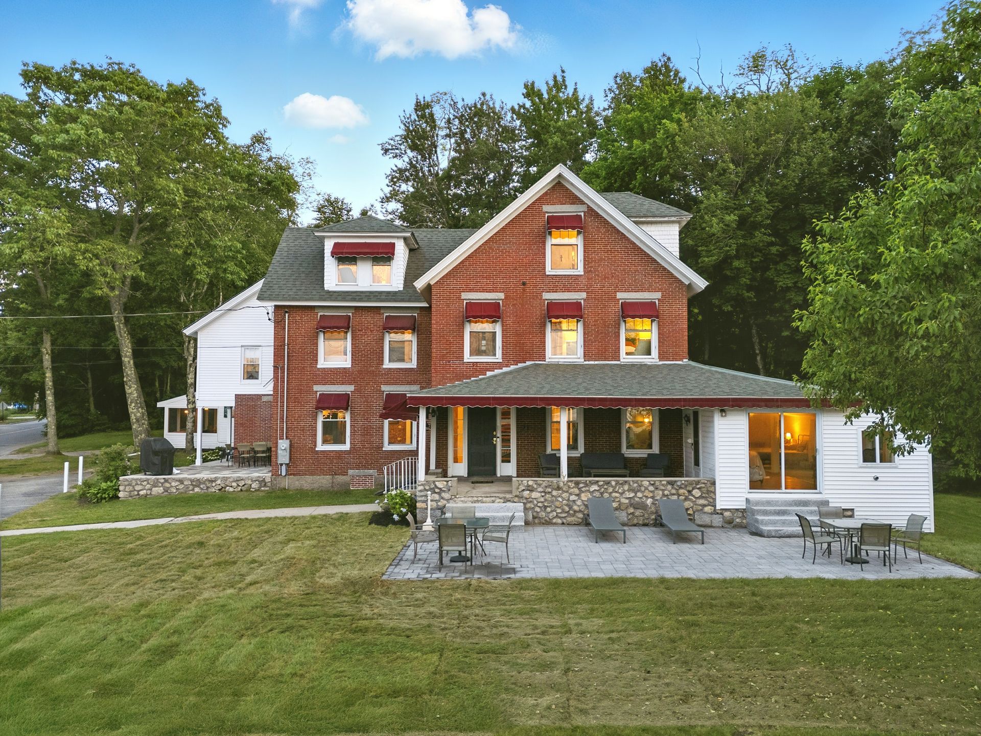 Two-story red brick house with porch and awning. Lawn in front with patio furniture; trees in the background.