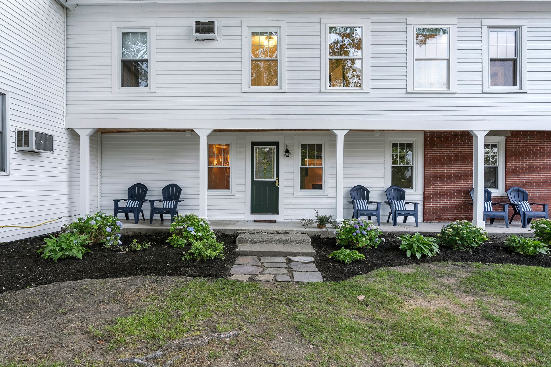 White two-story house with porch and blue chairs. Green plants and dark mulch in front.