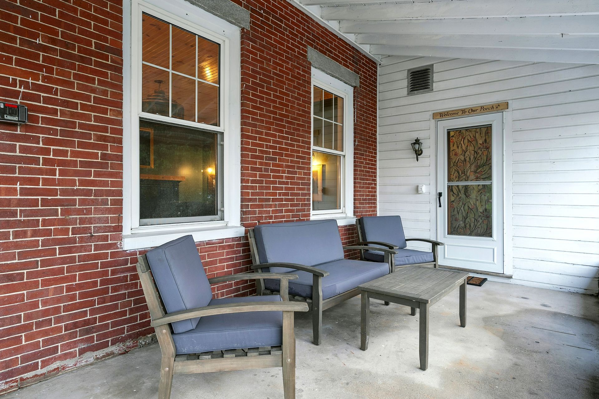 Covered porch with brick wall, two windows, gray furniture, and a door.