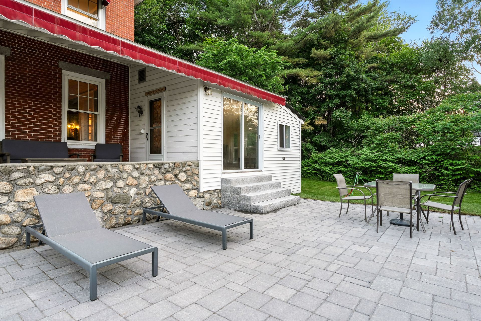 Patio with lounge chairs, table, and chairs outside a brick and white building with greenery.